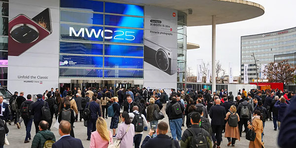 A large crowd gathers outside a modern convention center with glass walls and a sign reading “MWC25.” Two large banners display foldable smartphones. Office buildings and leafless trees are in the background, suggesting a tech event in a business district.