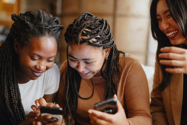 Three women smiling and laughing together whilst looking at their mobile phones indoors.